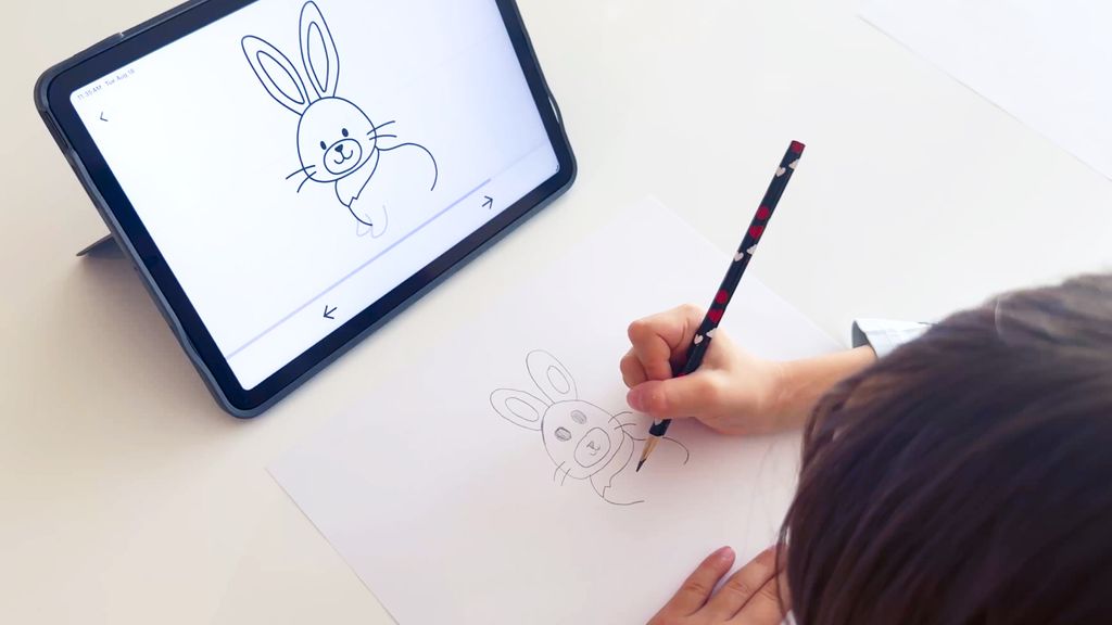 Child drawing on paper with a pencil while following a step-by-step drawing guide on a tablet