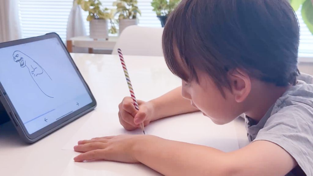Child drawing on paper with a pencil while following a step-by-step guide on a tablet