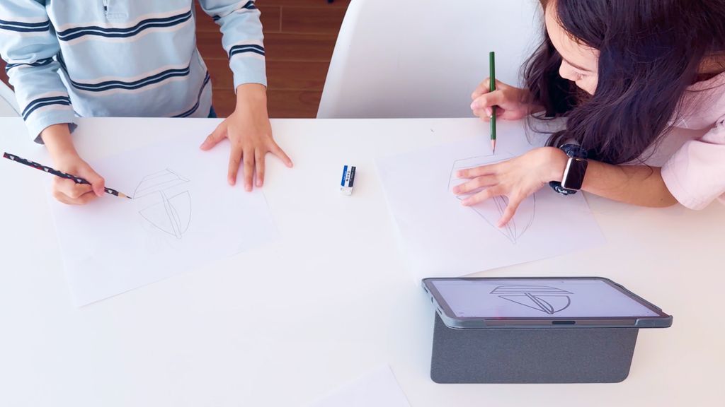 Two children drawing together on paper while following a drawing guide on a tablet